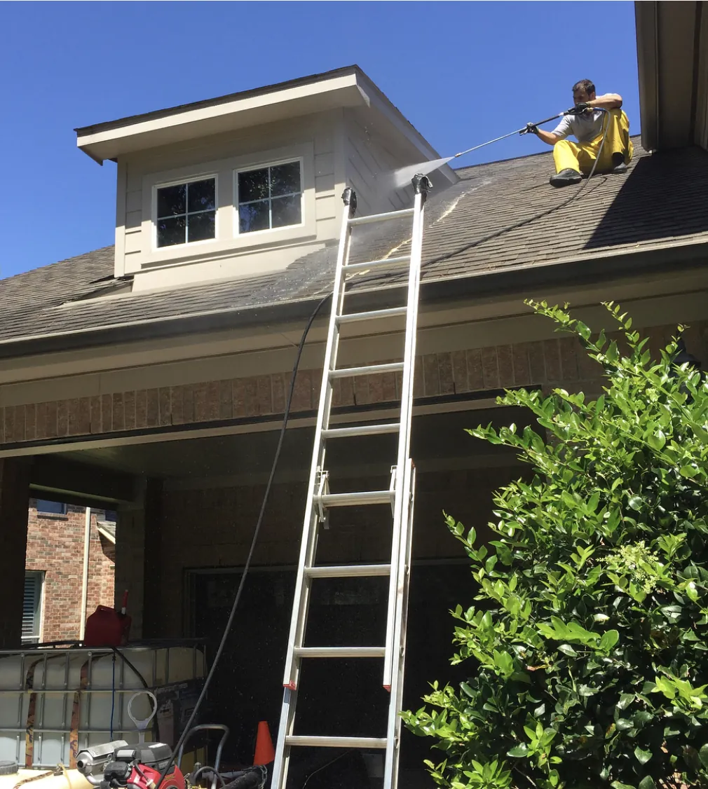 Mr. Power Wash technician on a shingle roof soft washing it with a spray wand, with a ladder and equipment below.